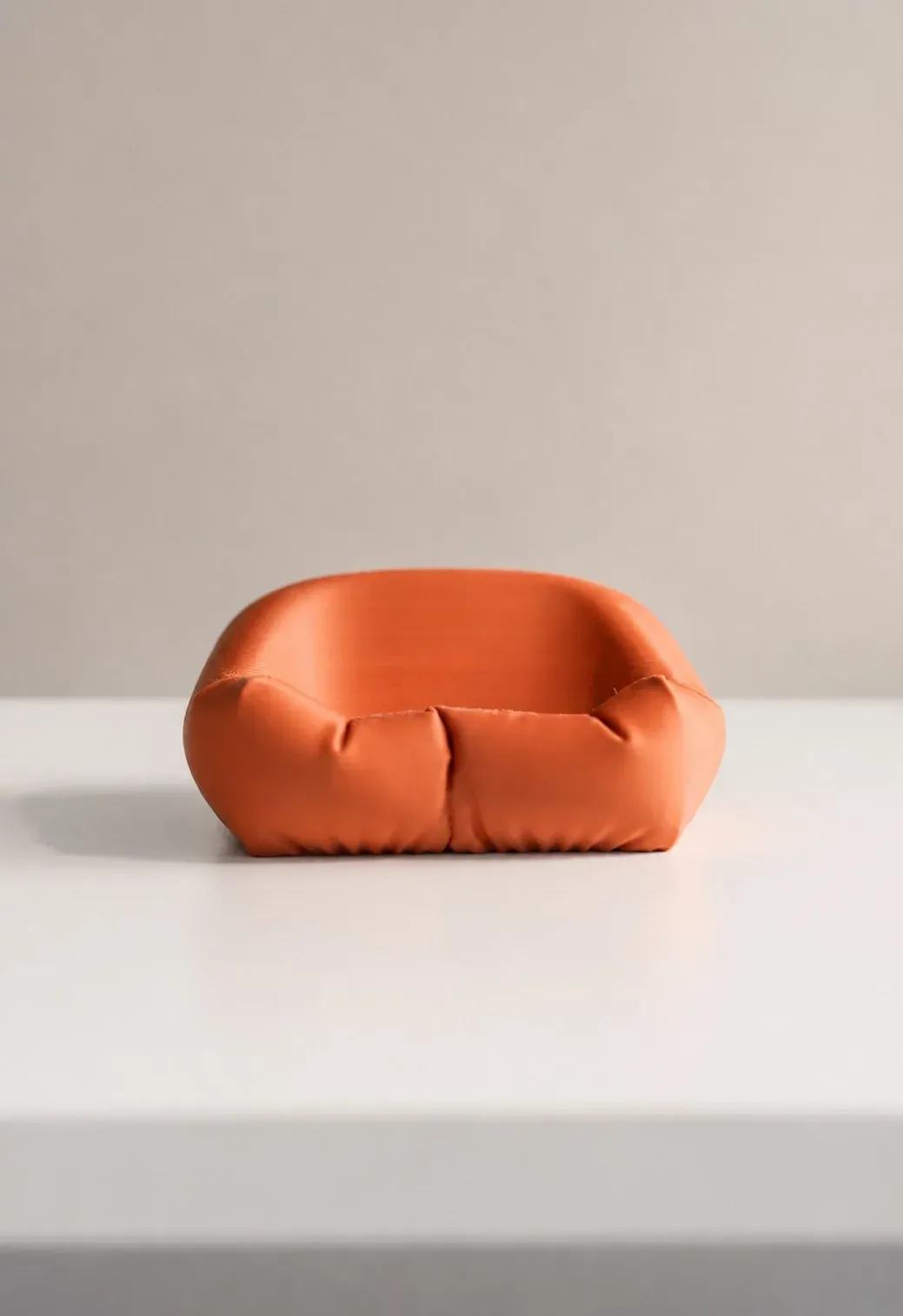 Cama cojín naranja impresa en 3D, vacía y de frente sobre una mesa blanca